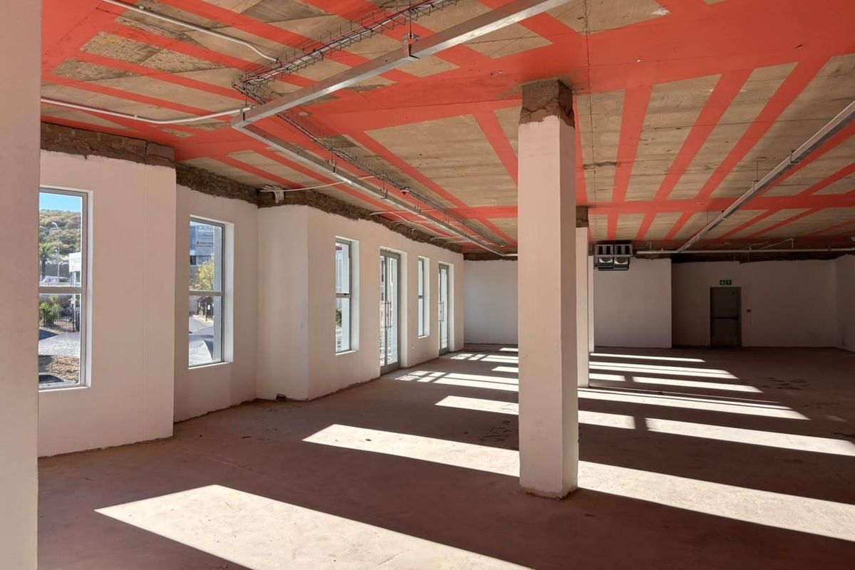 An open-plan office space in shell condition, featuring exposed concrete ceilings, structural columns, and large windows providing ample natural light.
