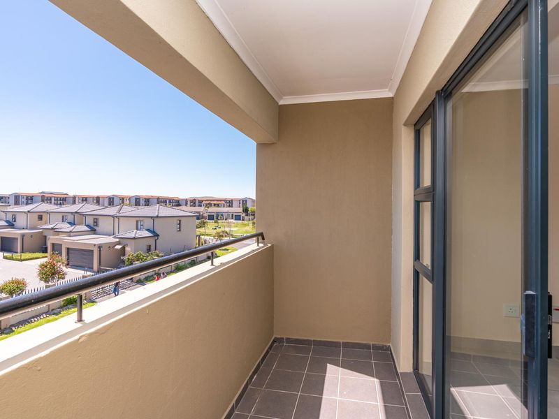 A clean, modern balcony featuring tiled flooring, a metal balustrade, and a sliding glass door, offering an open view of the surrounding residential estate.
