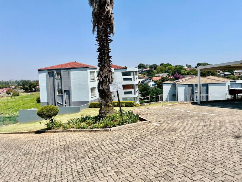 Exterior view of the apartment complex featuring a paved driveway, carport, and well-maintained communal garden areas under a clear blue sky.