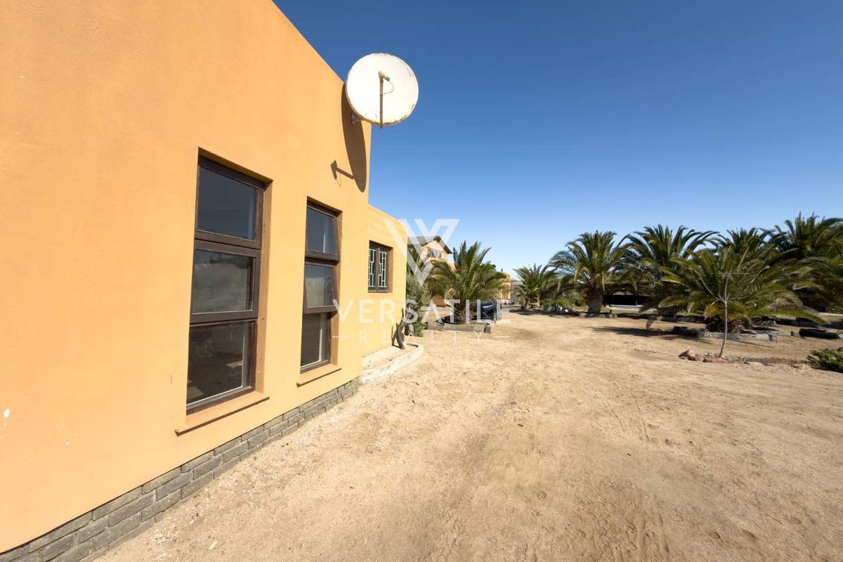 Rear exterior view of the property featuring a plastered wall, aluminium window frames, and a spacious, undeveloped sandy yard with mature palm trees.