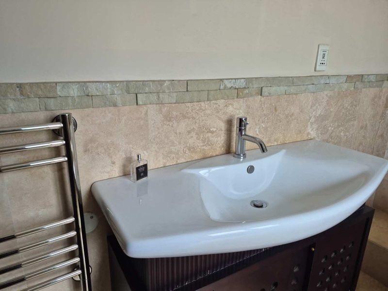 A bathroom vanity area featuring a white basin, chrome mixer tap, heated towel rail, and decorative travertine wall tiling.