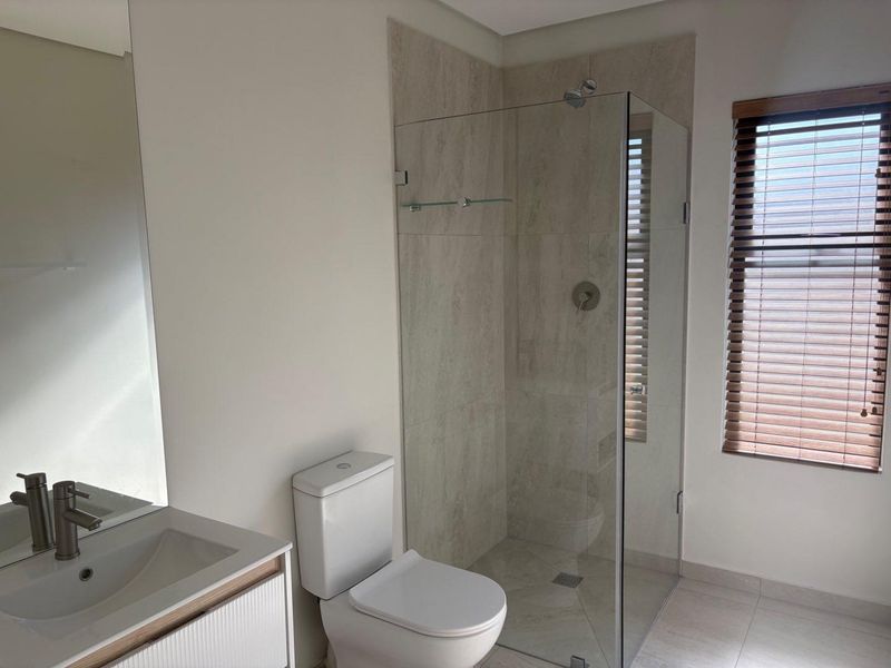 Modern bathroom featuring a walk-in glass shower, contemporary vanity with basin, and a toilet, finished with neutral floor-to-ceiling tiling and wooden blinds.