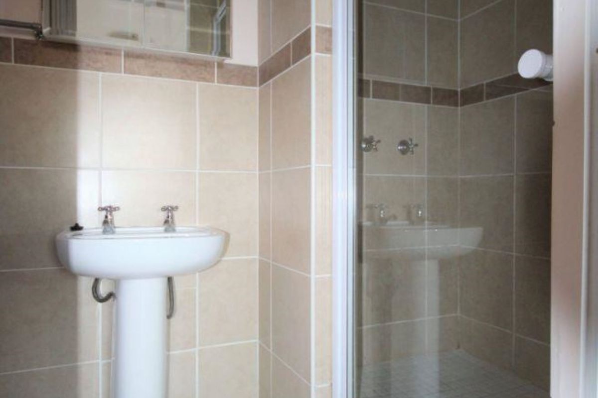 A functional bathroom featuring a pedestal basin, a glass-enclosed shower, and neutral wall and floor tiling.