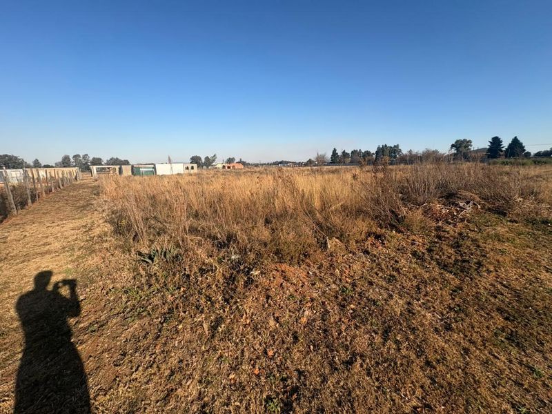A wide-angle view of a vacant residential plot featuring dry, overgrown grass and perimeter wire fencing under a clear blue sky.