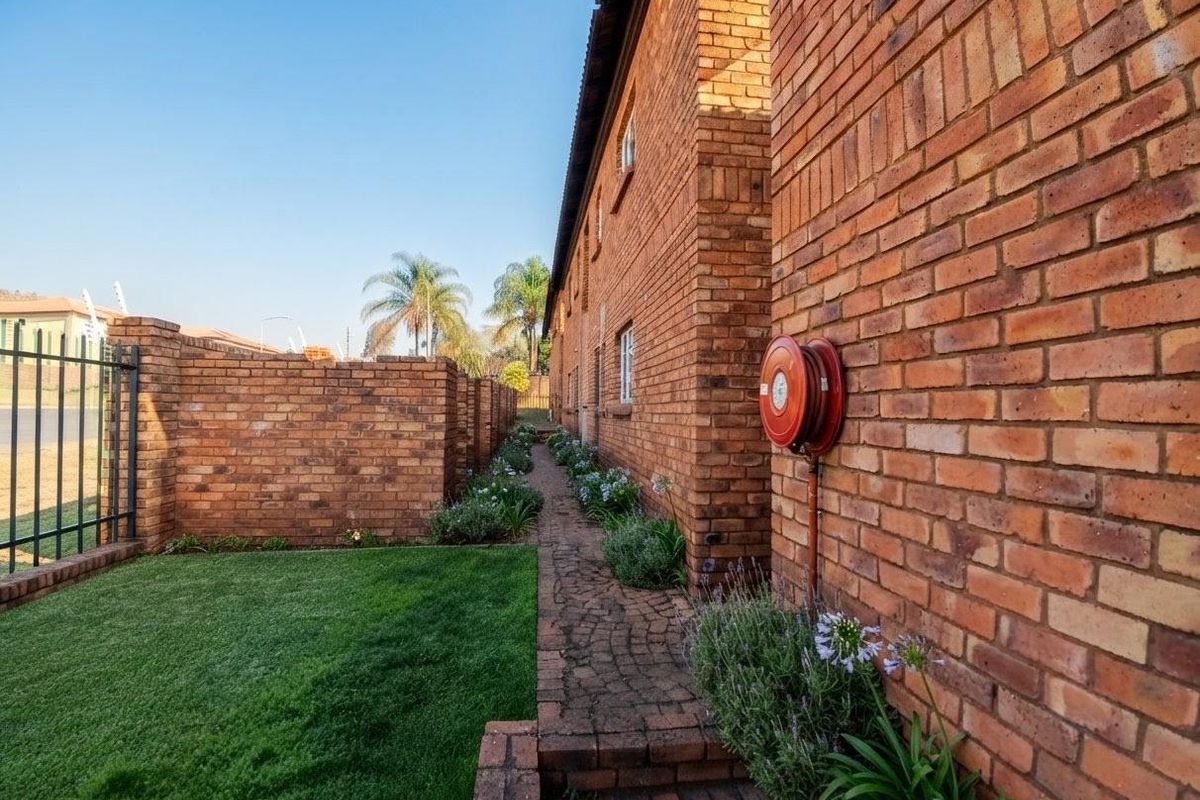 A well-maintained exterior side passage featuring face brick walls, a paved walkway, a neat lawn, and a wall-mounted fire hose reel.