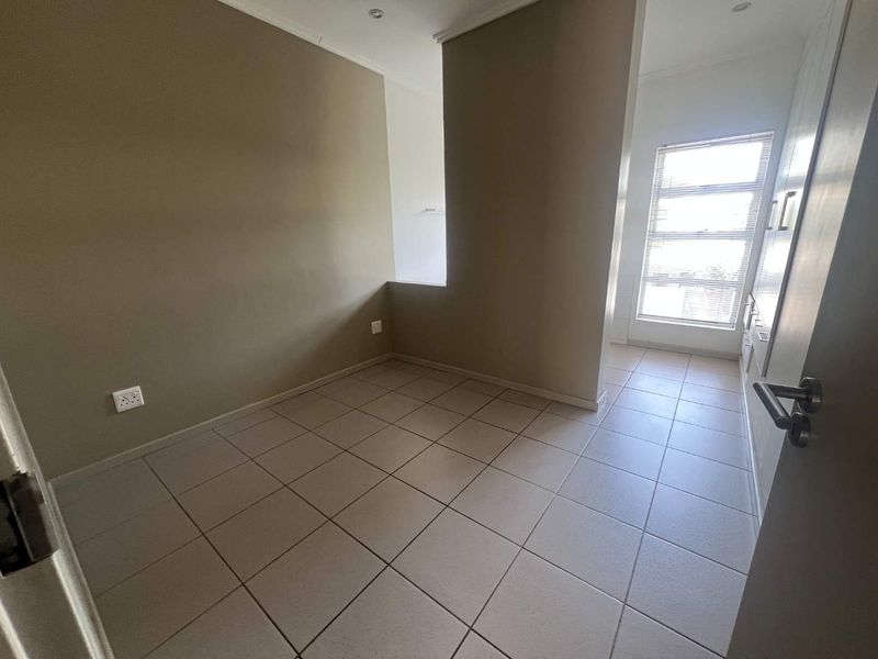 An empty, neutrally painted bedroom featuring tiled flooring, recessed lighting, and a window with fitted blinds, offering a clean and versatile living space.