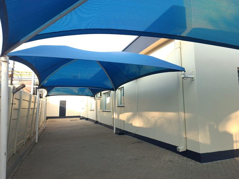 A paved outdoor area featuring a large blue shade sail structure, providing covered parking or utility space alongside the office building.