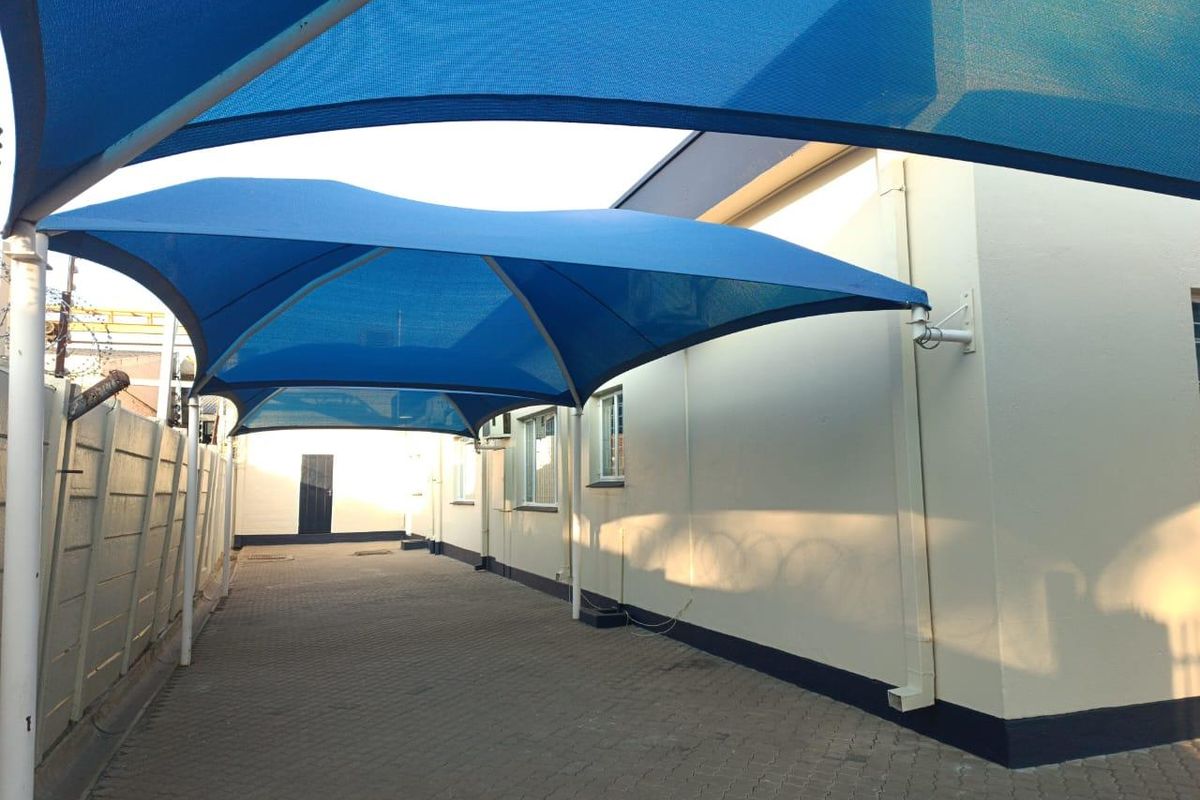 A paved outdoor area featuring a large blue shade sail structure, providing covered parking or utility space alongside the office building.