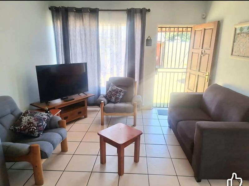 A functional living room featuring tiled flooring, natural light through large windows, and direct access to the exterior via a secure timber door.