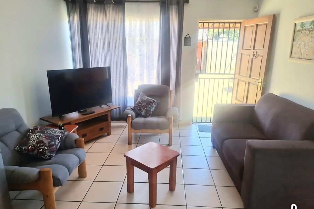A functional living room featuring tiled flooring, natural light through large windows, and direct access to the exterior via a secure timber door.