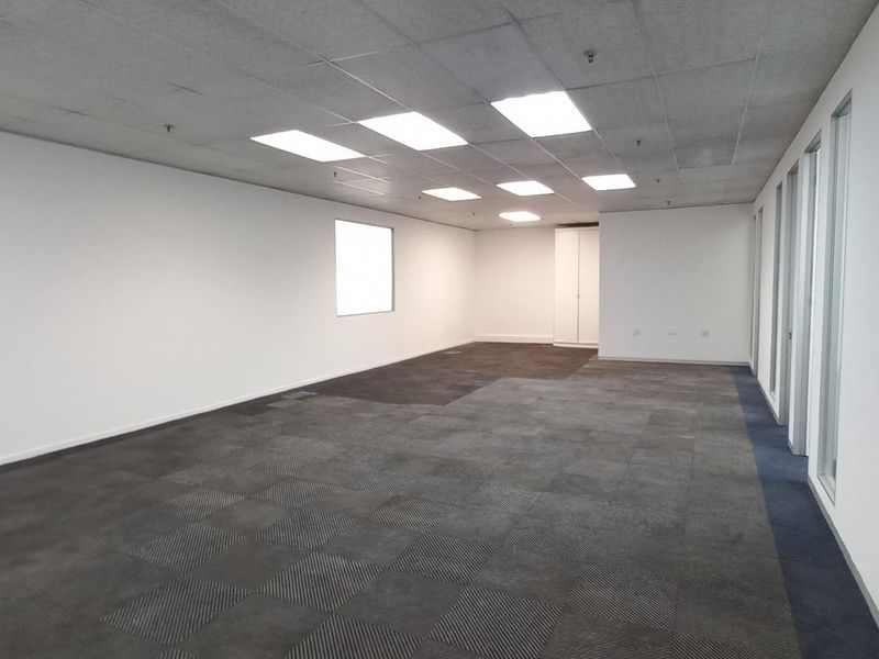 A spacious, empty open-plan office area featuring neutral walls, carpet tile flooring, and a suspended ceiling with recessed lighting.