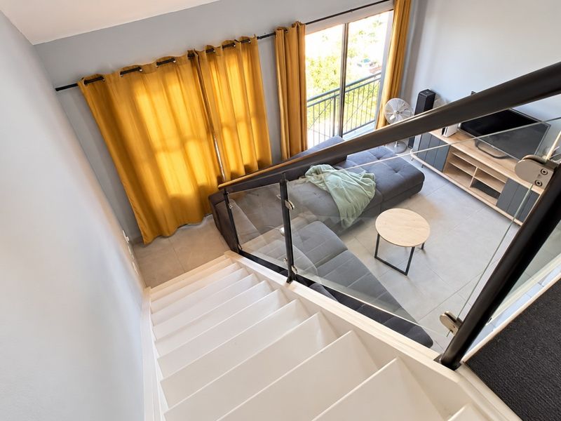 A bright living room viewed from the staircase, featuring modern glass balustrades, tiled flooring, and a sliding door leading to a balcony.