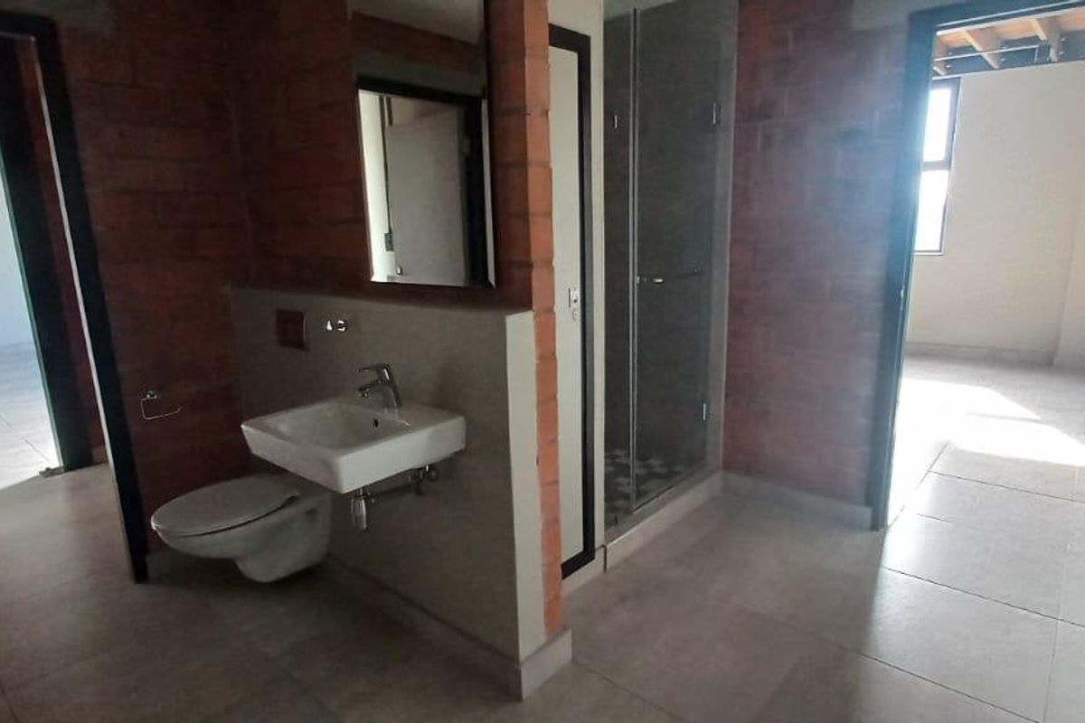 A modern industrial-style bathroom featuring a wall-mounted basin, toilet, and glass-enclosed shower, set against exposed brick walls with tiled flooring.