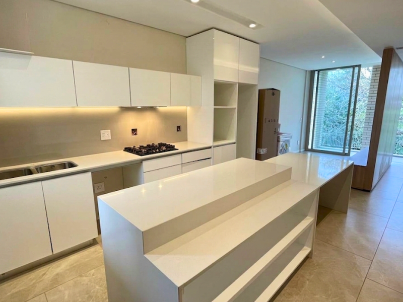 Modern kitchen featuring sleek white cabinetry, stone countertops, a central island, and integrated gas hob, flowing into an open-plan living area with balcony access.