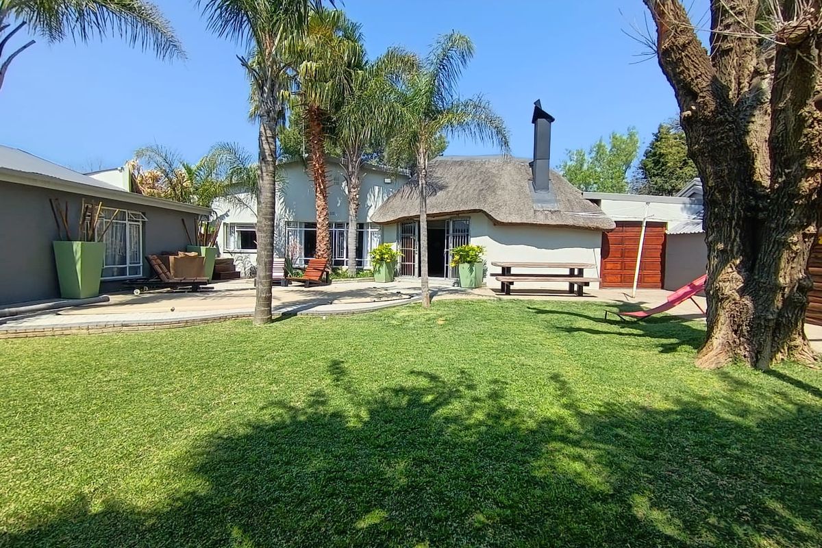 A spacious rear garden featuring a well-maintained lawn, paved patio area, and a distinctive thatched-roof structure with an external chimney.