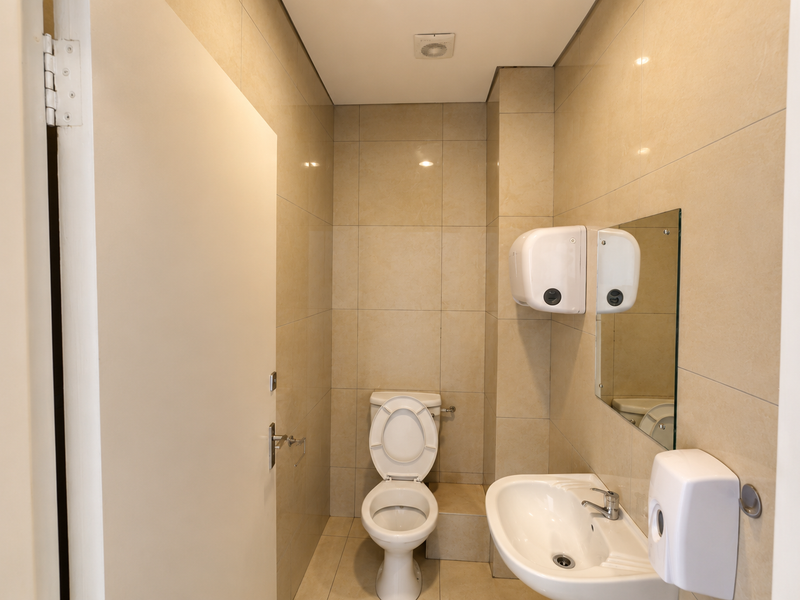 A clean, modern office restroom featuring neutral wall tiles, a white toilet, a wall-mounted basin, and essential sanitary fixtures.