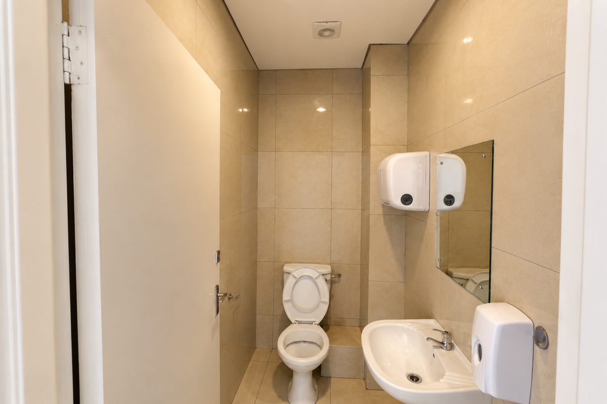 A clean, modern office restroom featuring neutral wall tiles, a white toilet, a wall-mounted basin, and essential sanitary fixtures.