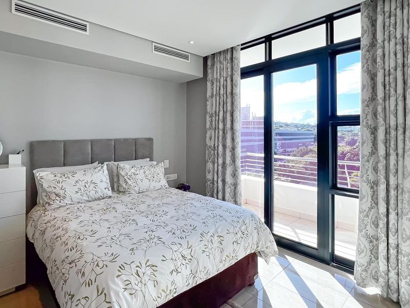 A bright bedroom featuring floor-to-ceiling glass doors leading to a private balcony, integrated air conditioning, and neutral finishes.