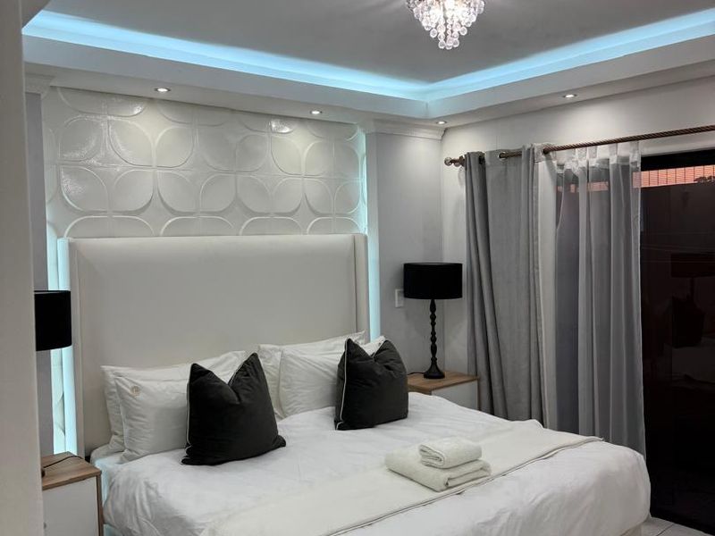 Modern bedroom featuring a textured feature wall, recessed ceiling with ambient cove lighting, and polished tiled flooring with access to an outdoor area.