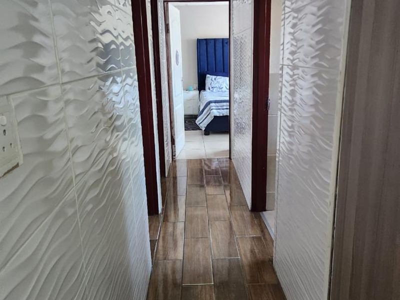 A narrow hallway featuring textured white wall tiles, wood-look floor tiles, and a glossy ceiling finish, leading towards a bedroom.