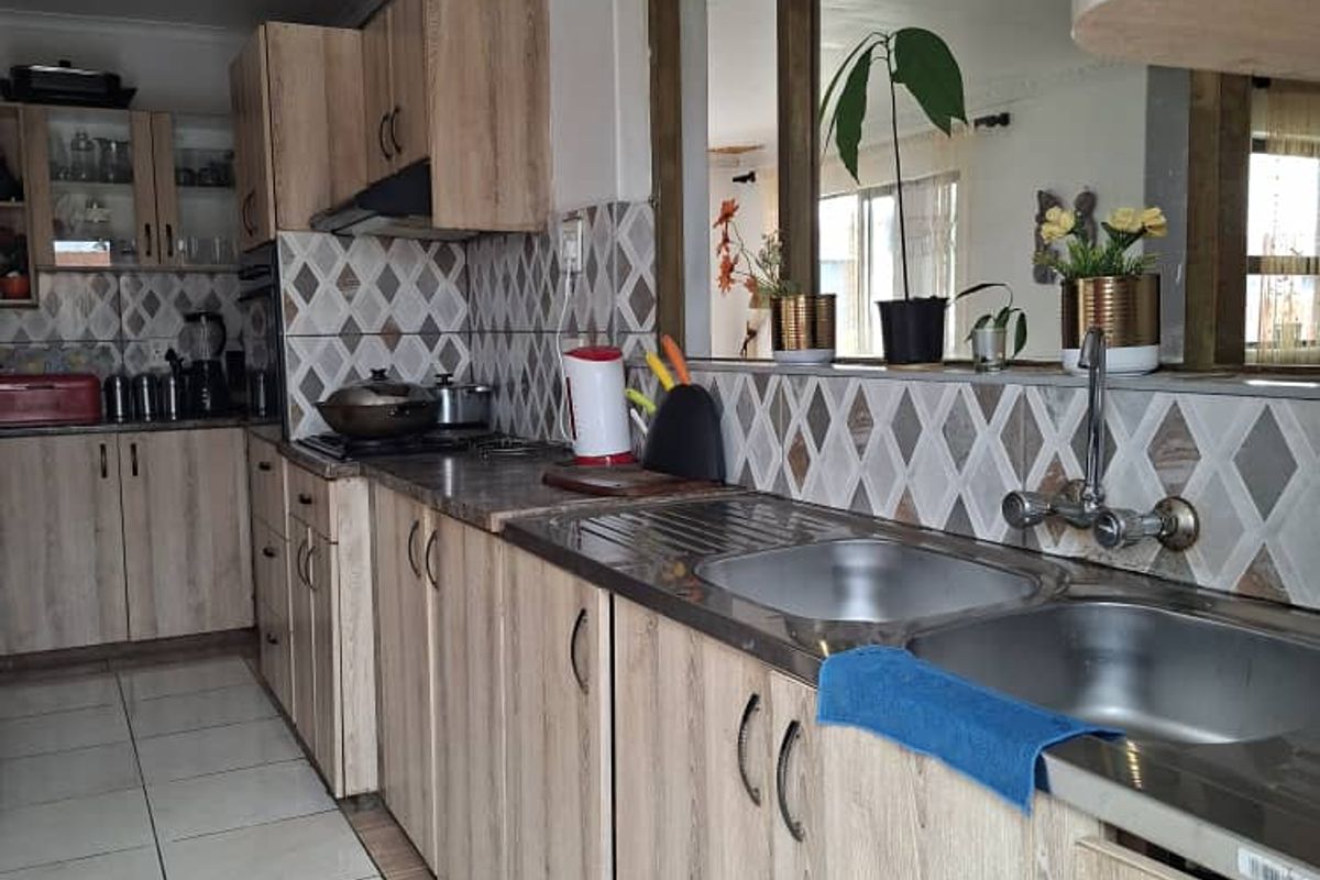 A functional kitchen featuring light wood-grain cabinetry, patterned tile backsplash, double stainless steel sink, and tiled flooring with a pass-through window to the living area.