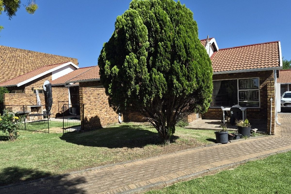 A view of the rear exterior of the apartment, featuring a brick facade, tiled roof, paved walkway, and a well-maintained garden area.