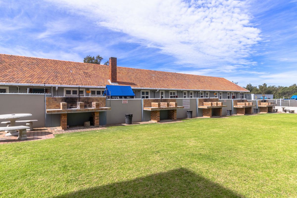 Communal outdoor area featuring a row of built-in brick braais, a well-maintained lawn, and a long residential building with a tiled roof.