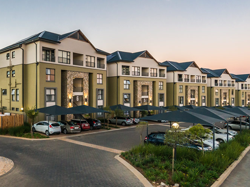 A modern apartment complex featuring stone-clad facades, private balconies, and extensive paved parking areas with shade cloth carports.