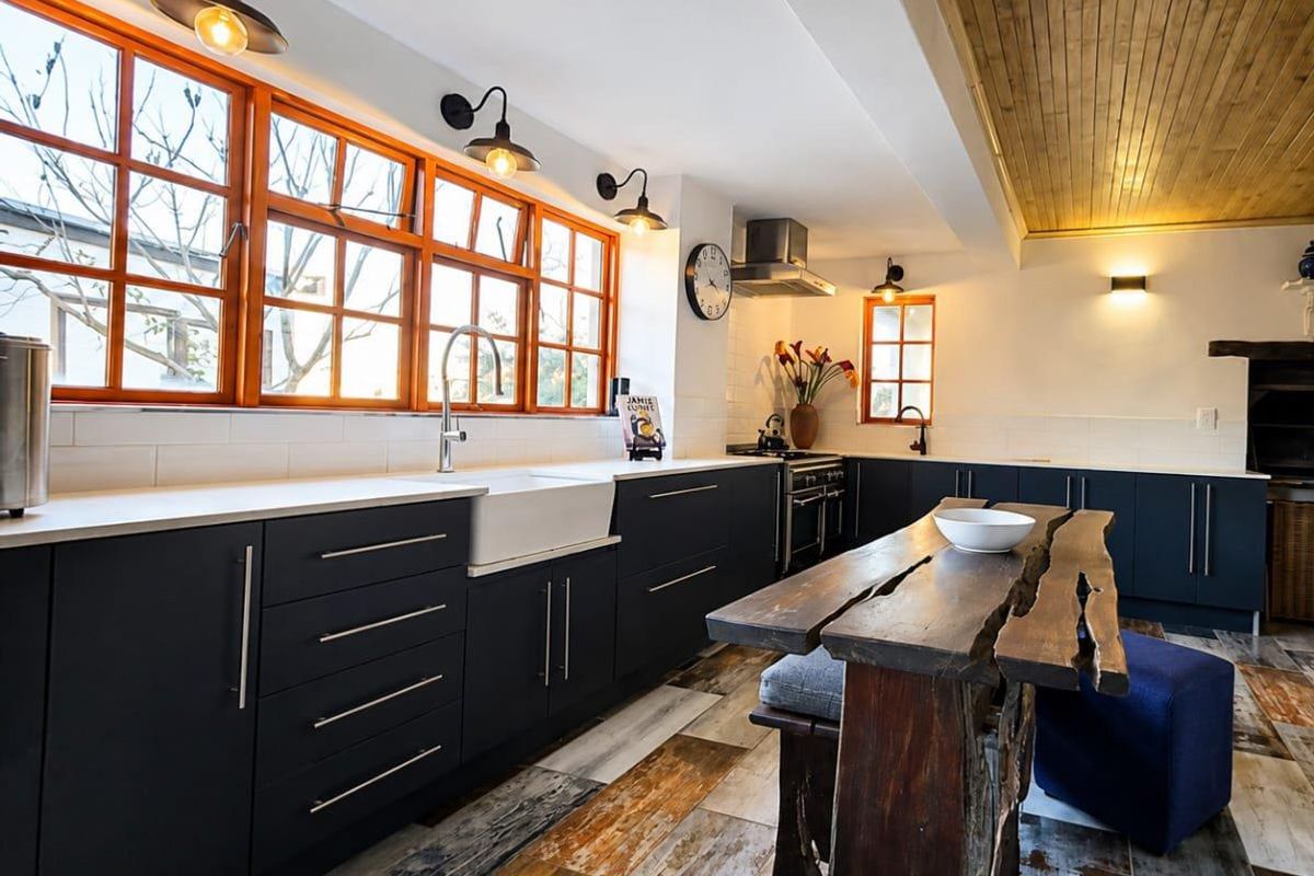 A modern kitchen featuring dark cabinetry, a farmhouse sink, gas stove, extractor fan, and unique timber ceiling accents with warm lighting.