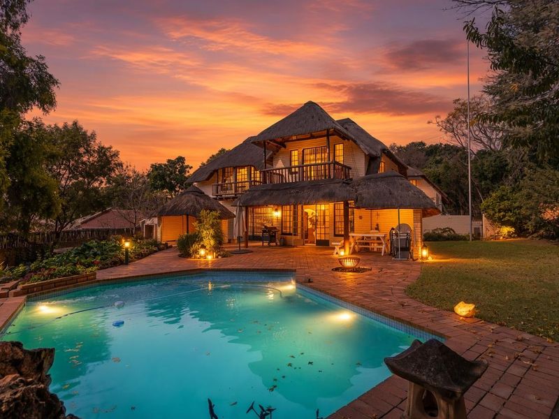A beautiful rear exterior view of a thatched-roof house featuring a swimming pool, brick-paved patio area, and lush garden at sunset.