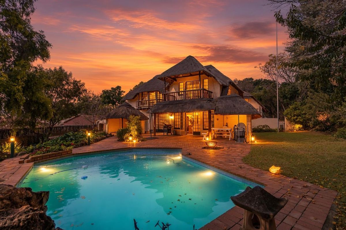 A beautiful rear exterior view of a thatched-roof house featuring a swimming pool, brick-paved patio area, and lush garden at sunset.