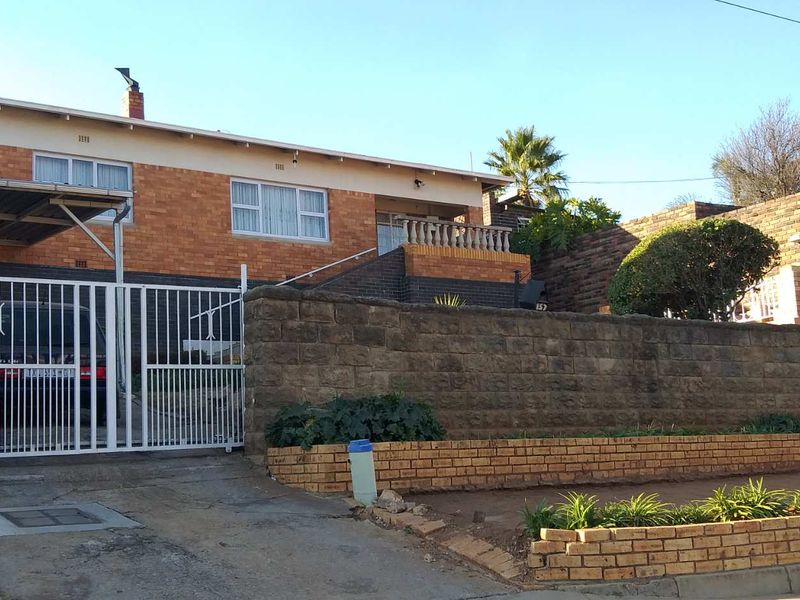 Front elevation of a brick house featuring a carport, secure gated entrance, and tiered retaining walls with established garden beds.