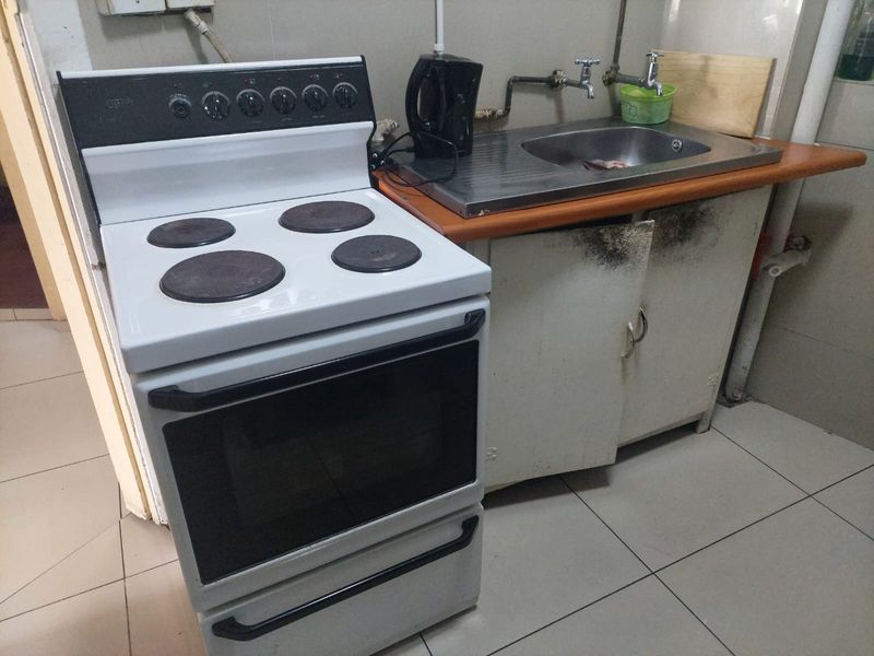 A compact kitchen area featuring a freestanding stove, stainless steel sink, and tiled flooring, currently showing significant wear and damage to the cabinetry.