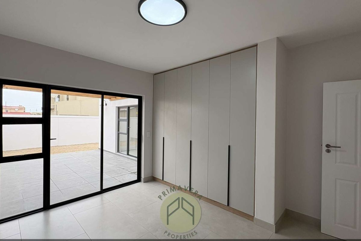 A bright, modern bedroom featuring neutral tiled flooring, floor-to-ceiling built-in cupboards, and a large sliding glass door providing access to the patio.