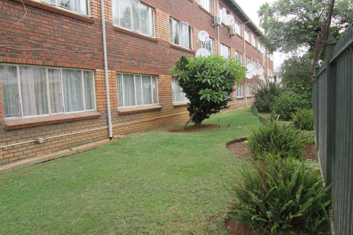 Exterior view of the apartment building featuring a brick facade, ground-level windows, and a shared garden area with established landscaping.