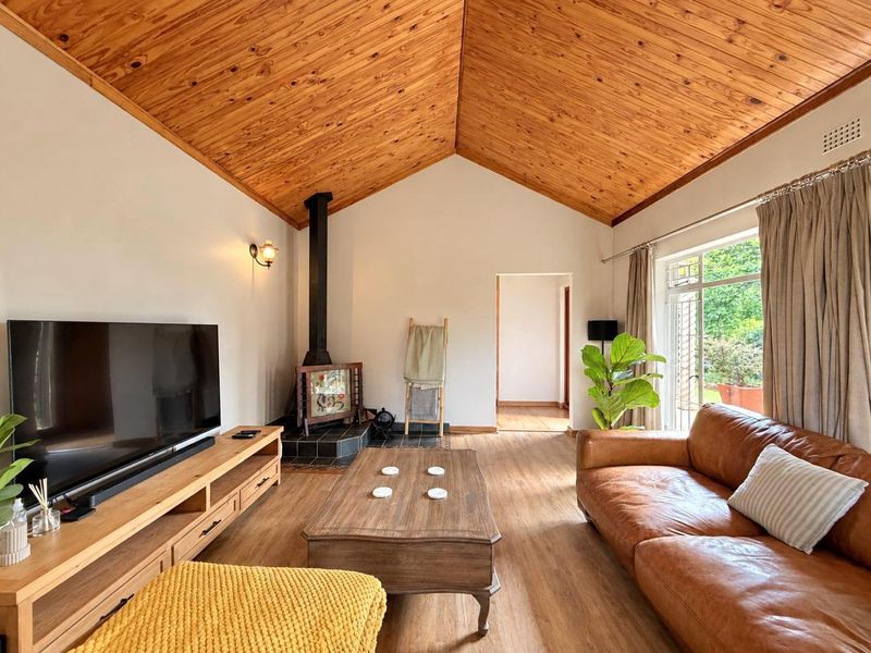 A cozy living room featuring a vaulted timber ceiling, a freestanding wood-burning fireplace on a tiled hearth, and large windows overlooking the garden.