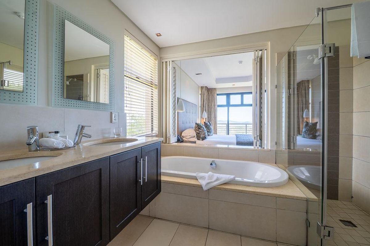 Modern en-suite bathroom featuring a double vanity, built-in bathtub, and glass-enclosed shower, with folding doors opening into the adjacent bedroom.