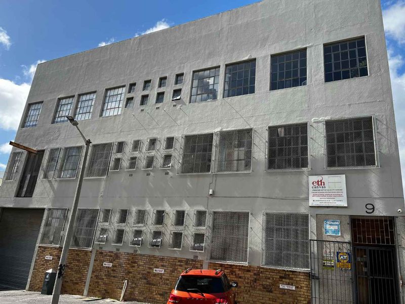 Three-story industrial building facade featuring steel-framed windows, security grilles, and a ground-level roller shutter door entrance.