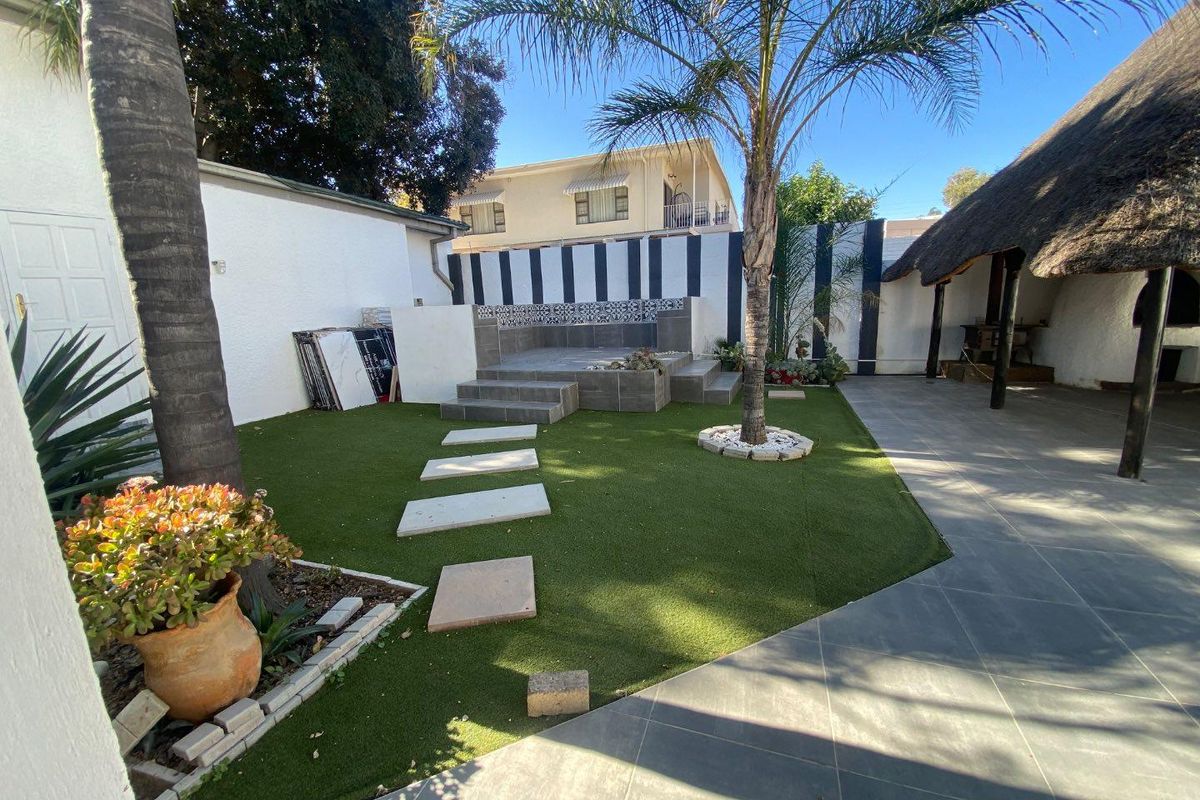 A landscaped rear garden featuring artificial turf, stone stepping paths, a raised tiled seating area, and a covered thatched patio space.