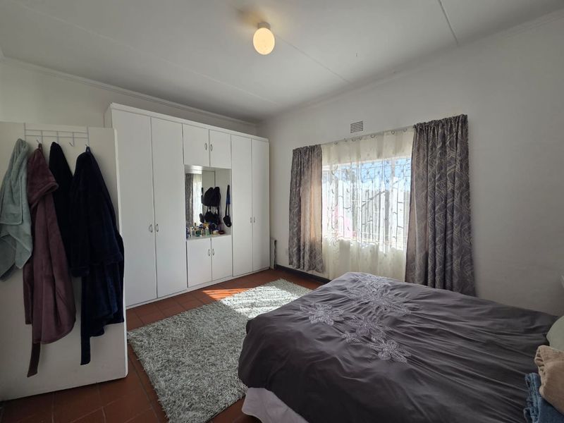 A bedroom featuring built-in cupboards with a central mirror, tiled flooring, and a window fitted with security bars.