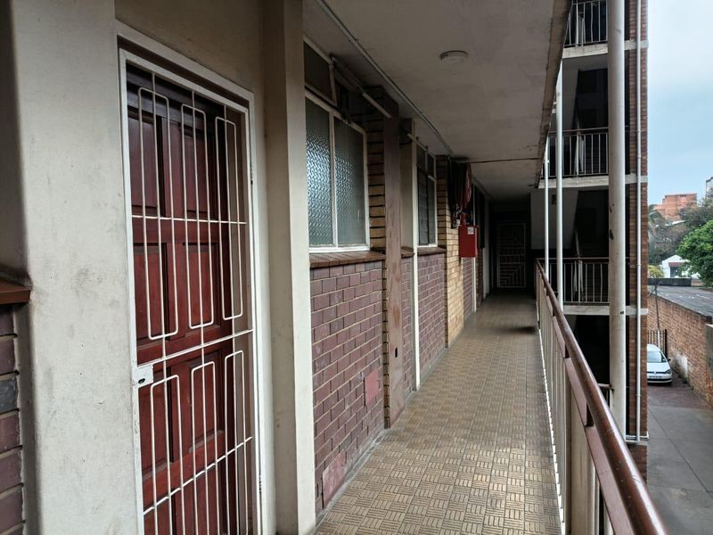 An exterior apartment building walkway featuring brick facade walls, tiled flooring, and metal safety railings with a security gate entrance.
