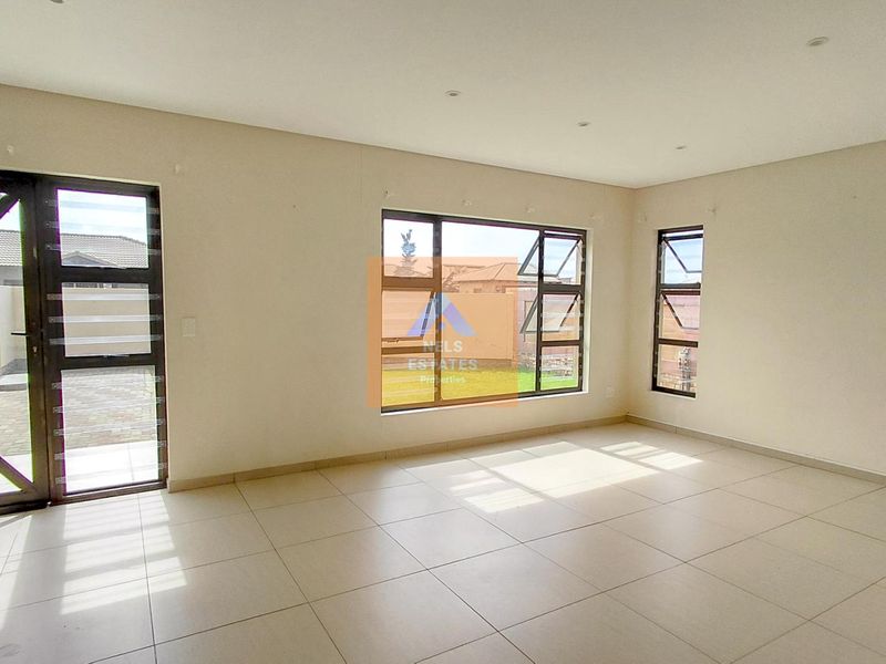A bright, empty living area featuring neutral tiled flooring, modern aluminium window frames, and recessed downlighting, offering a clean canvas for interior design.