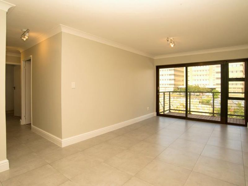 A bright, empty living area featuring neutral tiled flooring, modern ceiling spotlights, and a large sliding glass door leading to a private balcony.