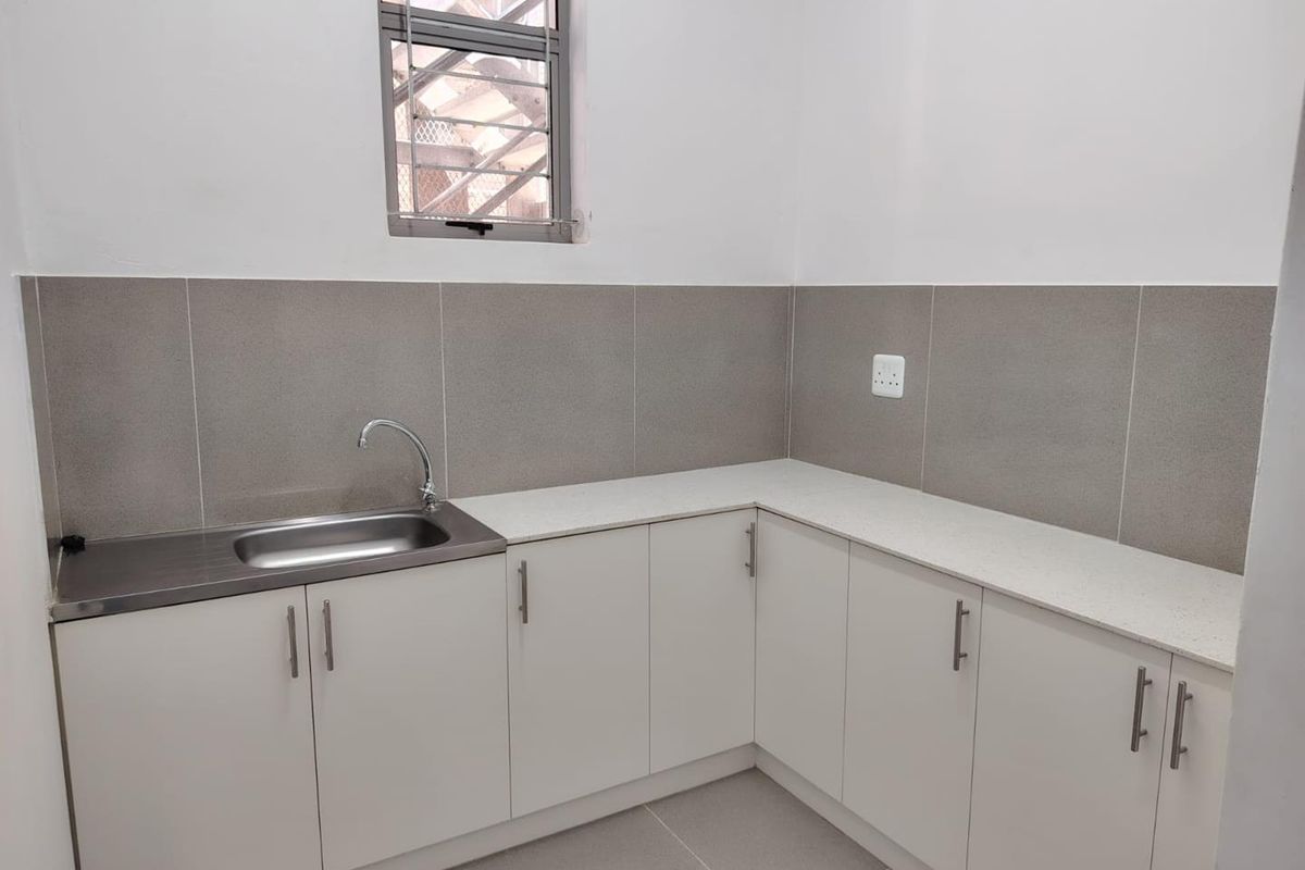 A clean, modern kitchenette area featuring white cabinetry, stone-look countertops, a stainless steel sink, and neutral tiled splashbacks.