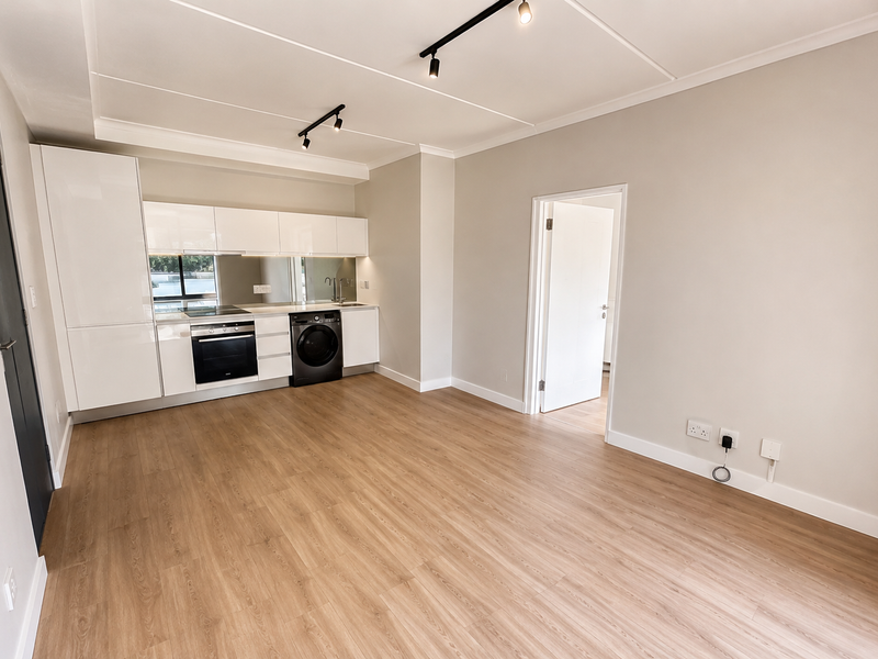 Modern open-plan living area featuring a sleek, white kitchen with integrated appliances, track lighting, and light wood-look laminate flooring throughout.