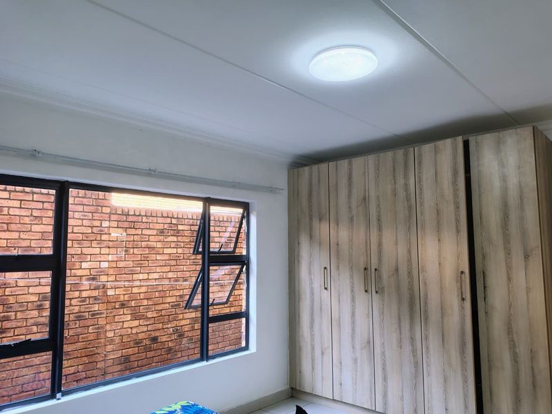 A bright bedroom featuring modern wood-finish built-in cupboards and large aluminium-framed windows overlooking a brick wall.