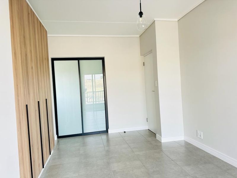 A bright, modern bedroom featuring neutral tiled flooring, built-in wood-finish cupboards with black handles, and a sliding glass door leading to a balcony.