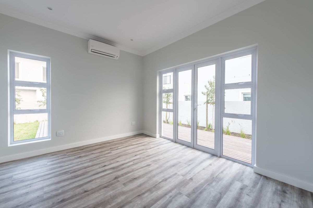 A bright, empty living room featuring modern wood-look flooring, neutral walls, a wall-mounted air conditioning unit, and large glass doors opening to a patio.