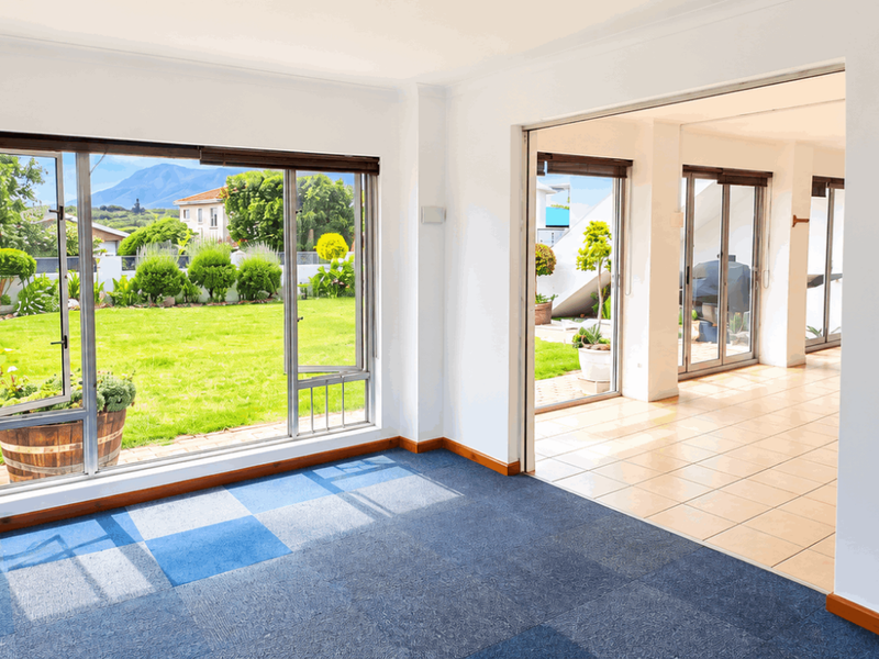 Bright and spacious living area featuring large windows with garden views, tiled and carpeted flooring, and seamless access to an adjoining patio space.