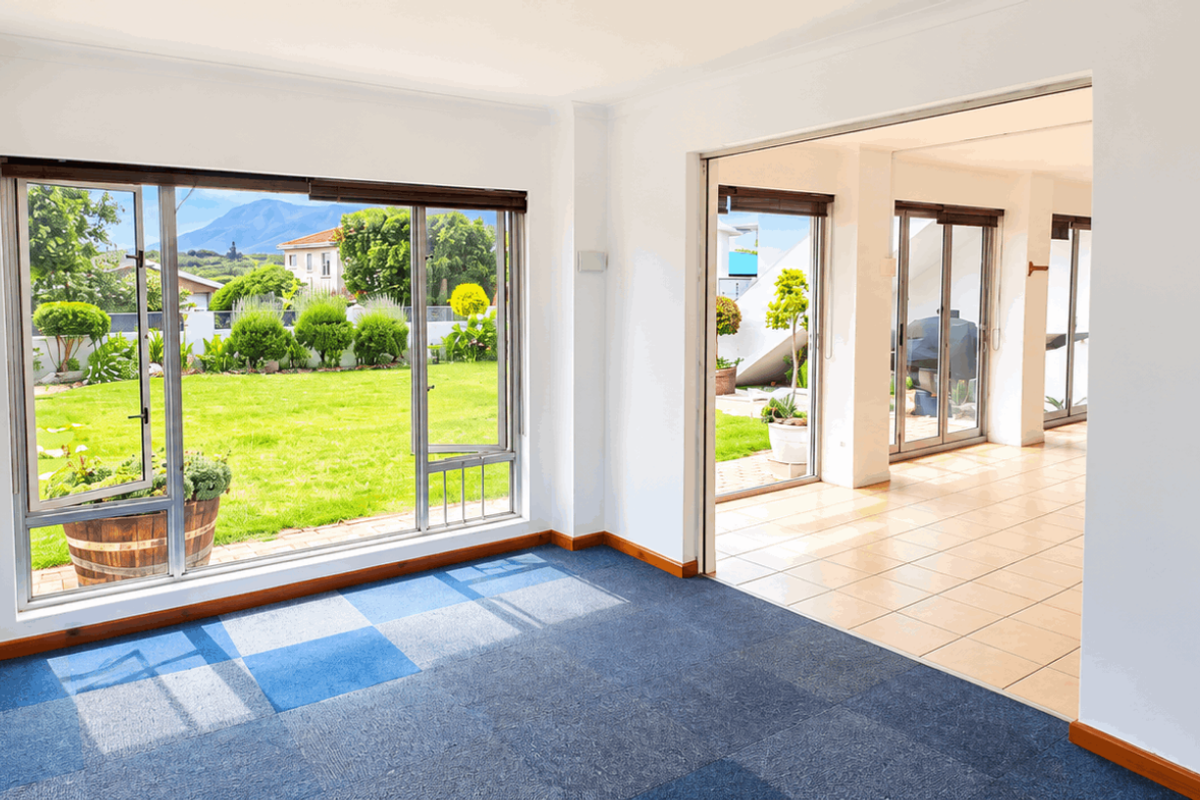 Bright and spacious living area featuring large windows with garden views, tiled and carpeted flooring, and seamless access to an adjoining patio space.
